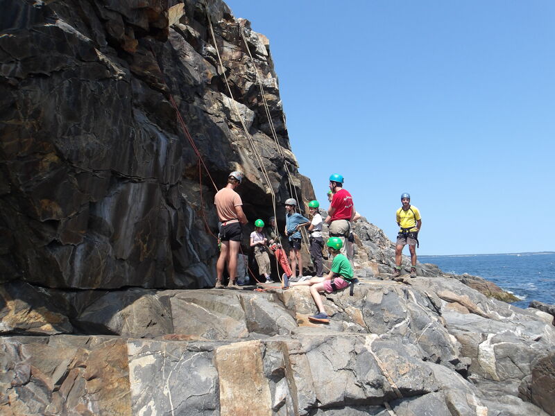 The image shows a group of people on a rocky cliff near the ocean. They appear to be involved in rock climbing or rappelling, as ropes are visible. Some individuals are wearing helmets, suggesting safety precautions. The weather seems sunny, and the overall scene depicts an outdoor adventure activity.
